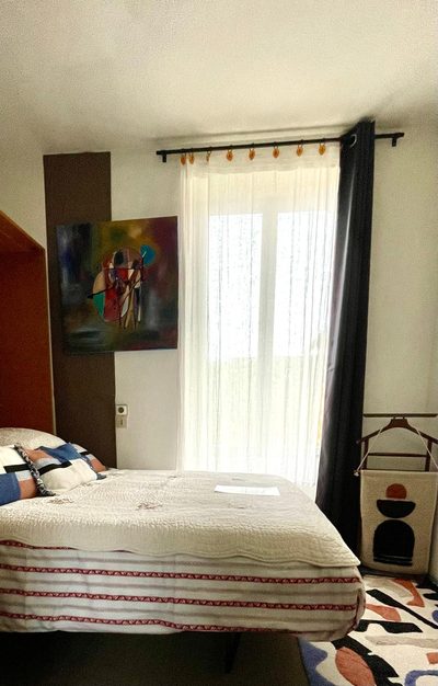 Chambre Brimbelle : lit double sous une baie vitrée voilée, tableau abstrait au mur et tapis à motifs au sol.