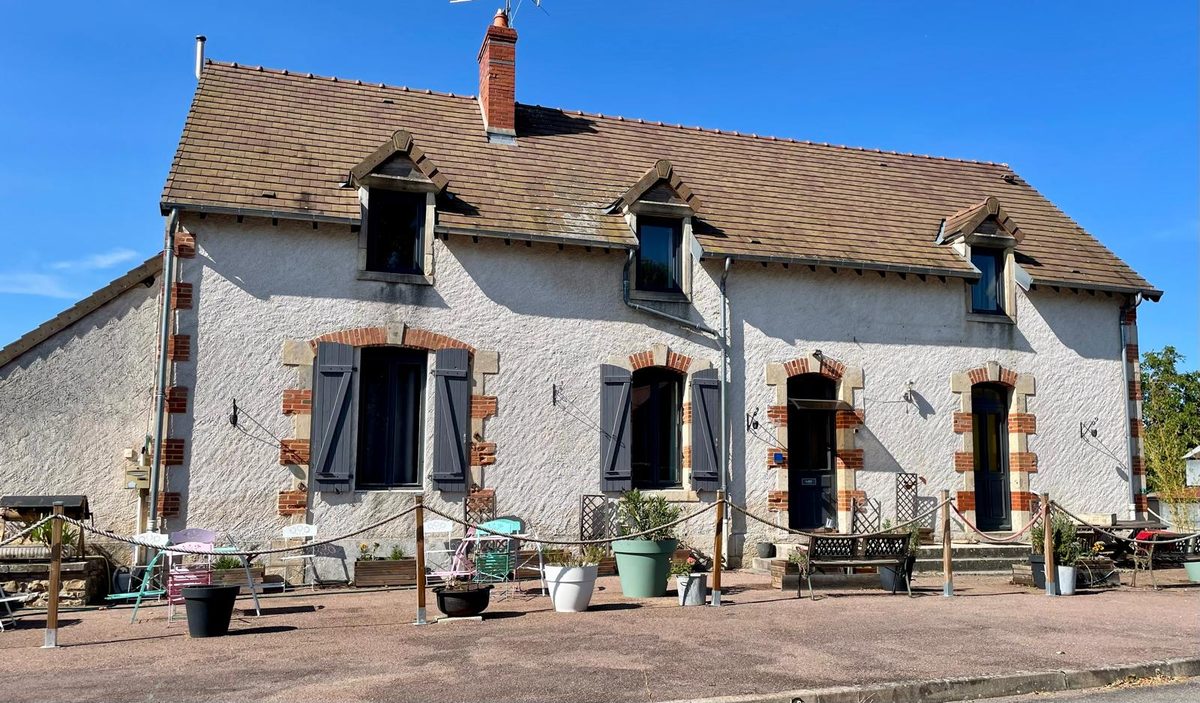 Façade de l'ancien relais : une longue maison enduite de blanc, encadrements de brique rouge, volets gris, trois lucarnes sur le toit de tuiles, tables et bancs alignés devant les portes.