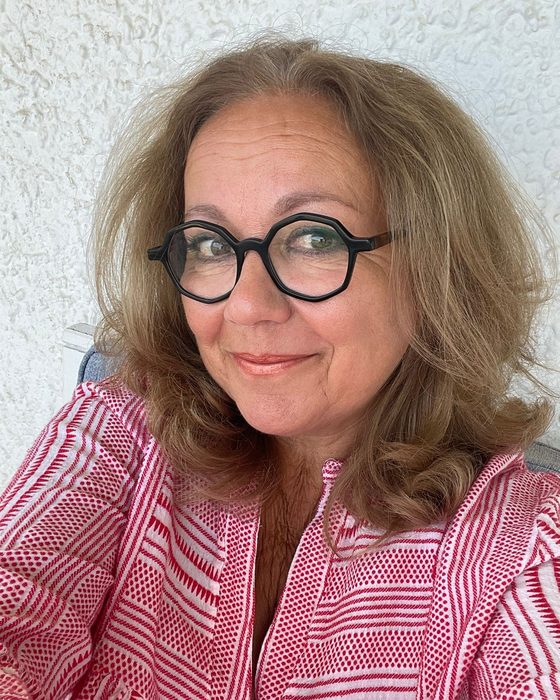 Sandrine James, cheveux blonds mi-longs et lunettes rondes noires, en tunique tissée rouge et blanche devant un mur crépi clair.
