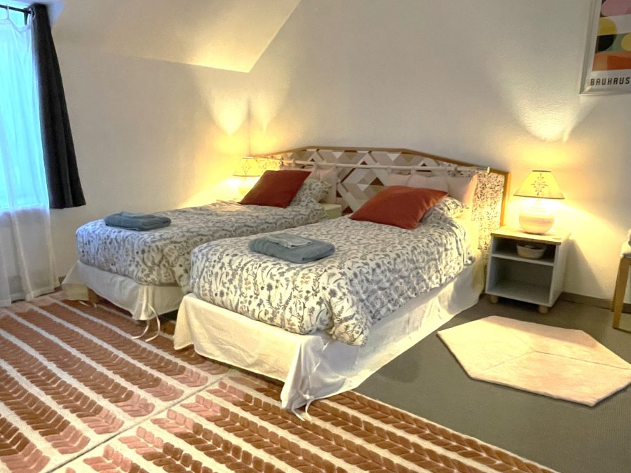 Chambre Bruyère : lit queen size, tête de lit en rotin, couvre-lit terre cuite, tapis tissé au sol.